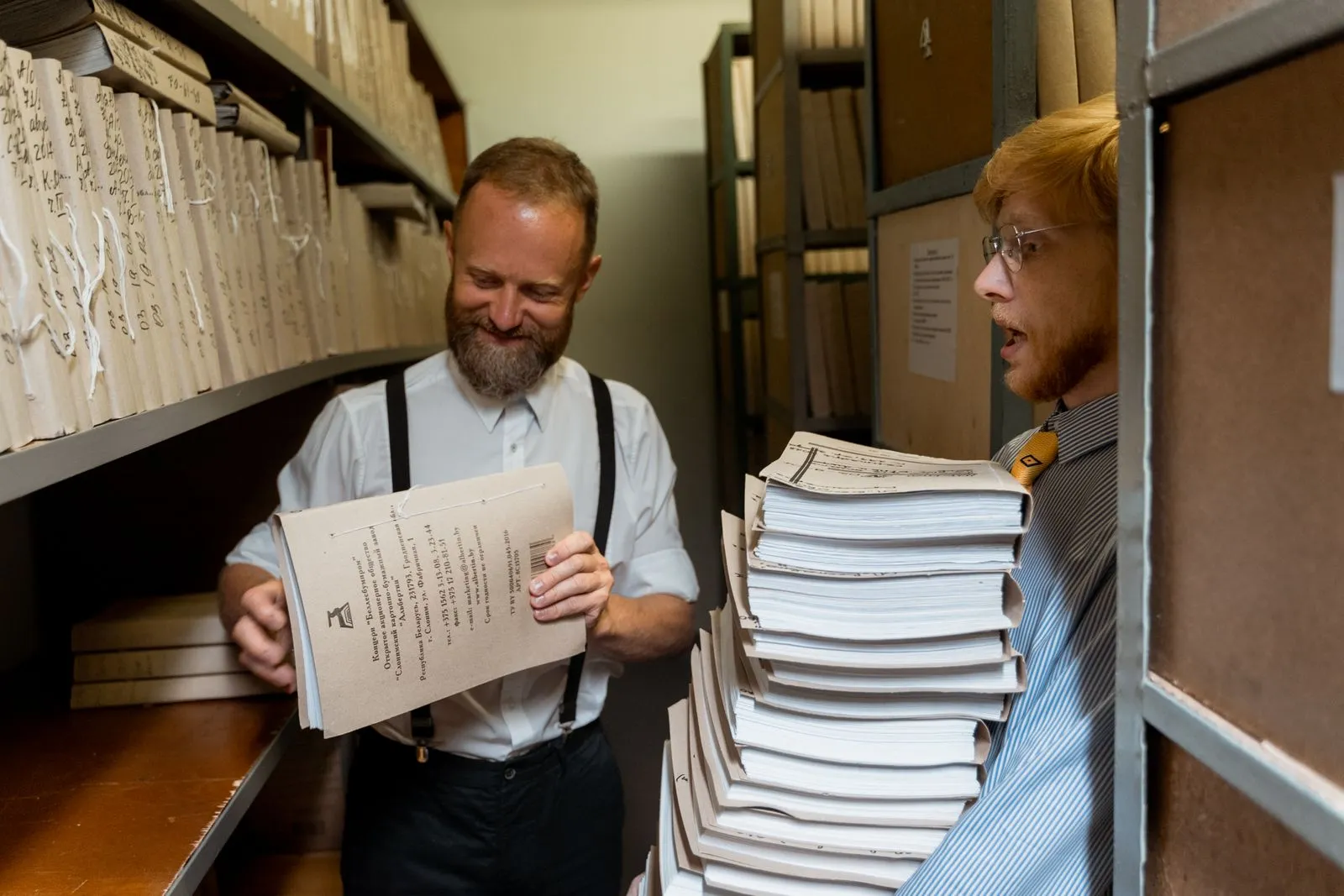Two men sorting through stacks of documents in an archive room, showcasing teamwork and organization.